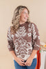 A woman wearing a brown and cream floral knit sweater with blue jeans, standing against a warm beige background.
