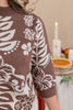 A close-up of a person wearing a brown and cream knit sweater with a botanical leaf and floral pattern, set against a softly blurred indoor background with a candle and pie.