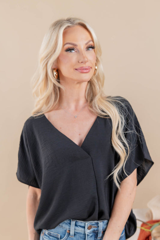 A blonde woman wearing a black short-sleeve wrap-style blouse with a V-neck and draped sleeves, paired with blue jeans, against a plain beige studio background.