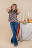 A woman posing in a brown and white plaid short-sleeve top and blue jeans, standing next to a decorated table with pumpkins, candles, and a pie against a solid beige background.