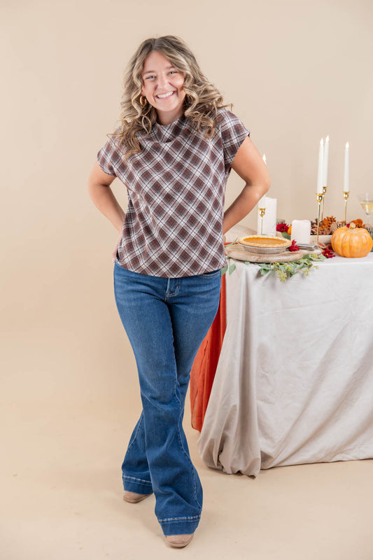 A woman modeling a brown and white plaid short-sleeve button-up shirt with blue jeans, standing in front of a beige studio backdrop next to a decorated autumn table.