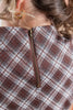 Close-up of the back of a plaid garment with a brown zipper, worn by a person with blonde hair and gold hoop earrings.