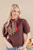 A smiling blonde woman wearing a brown short-sleeve blouse with puff sleeves and a leopard-print neck scarf tied at the collar, paired with blue jeans, against a warm beige studio background.