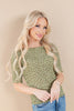 A blonde woman wearing a short-sleeve olive green knit top with a small brown floral pattern, paired with blue jeans, posing against a solid beige background.