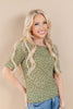 A smiling blonde woman wearing a short-sleeve olive green knit top with a brown dotted pattern, paired with blue jeans, posed against a solid beige studio background.