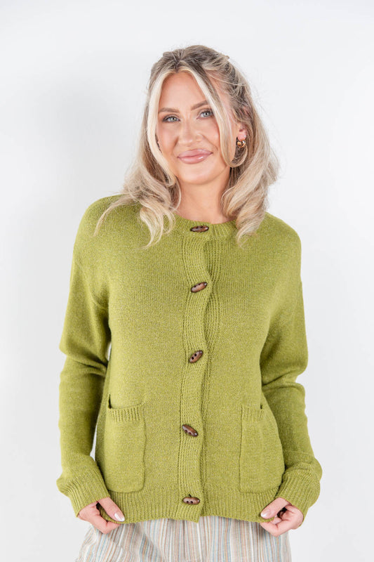 A woman modeling a light olive green knit cardigan sweater with brown toggle buttons, featuring a crew neck, front pockets, and ribbed cuffs and hem, set against a plain white studio background.