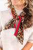 A close-up of a woman wearing a leopard print neck scarf with red and brown ribbon accents, styled tied around her neck over a cream ribbed top.