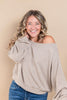A smiling woman with blonde-highlighted curly hair wearing a light taupe heather off-the-shoulder long-sleeve top and blue jeans, posed against a solid beige studio background.