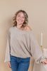 A woman smiling while wearing a light taupe heather off-the-shoulder long-sleeve sweater and blue jeans, standing against a solid beige background.