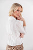 A blonde woman posing from a side-back angle wearing a white long-sleeve textured blouse with an adjustable back tie and leopard print pants against a plain white studio background.