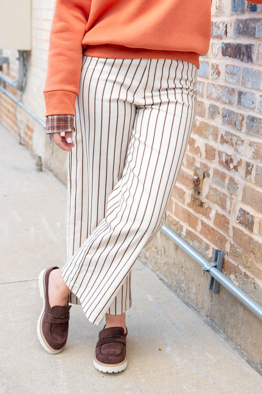 A person wearing cream wide-leg pinstriped pants with thin dark vertical stripes, an orange sweatshirt, and brown slip-on loafers, standing outdoors against a brick wall.