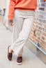 A person wearing cream wide-leg pinstriped pants with thin dark vertical stripes, an orange sweatshirt, and brown slip-on loafers, standing outdoors against a brick wall.