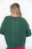 A woman viewed from behind wearing a dark green ribbed knit sweater with exposed seams, paired with red pants.