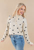 A smiling blonde woman wearing a cream knit sweater with black polka dots and light blue jeans, posed against a solid beige studio background.