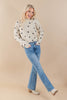 A woman modeling a cream-colored knit sweater with black polka dots, paired with light-wash blue jeans and beige ankle boots, against a solid beige studio background.