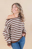 A woman wearing a brown and off-white horizontally striped off-the-shoulder knit sweater with blue jeans, posing against a solid beige studio background.