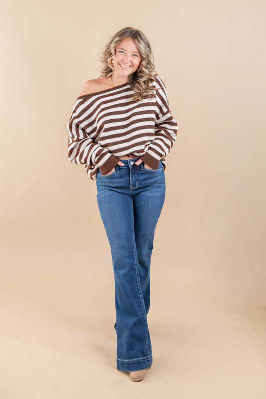 A woman modeling a brown and white striped off-the-shoulder long-sleeve top with blue flare jeans, standing against a solid beige studio backdrop.