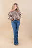 A woman modeling a brown and white striped off-the-shoulder long-sleeve top with blue flare jeans, standing against a solid beige studio backdrop.