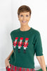 A woman wearing a dark green knit sweater with three embroidered nutcracker figures across the chest, paired with red and green plaid pants.