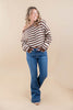 A woman modeling a brown and white horizontally striped off-the-shoulder long-sleeve top with blue jeans, standing against a solid beige studio backdrop.