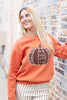 A woman wearing an orange crewneck sweatshirt with a plaid pumpkin graphic on the front, paired with cream and black pinstriped pants, standing against a brick wall.