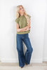 A blonde woman wearing a short-sleeve olive green mock-neck knit top and wide-leg blue jeans, posing against a white wall on a light wood floor.