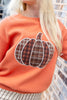A close-up of a person wearing an orange crewneck sweatshirt with a large applique pumpkin design on the front. The pumpkin pattern features brown, black, and white plaid sections outlined in white.