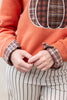 Close-up of a person wearing an orange sweatshirt with a plaid pumpkin graphic and matching plaid cuff details, paired with cream pinstriped pants.