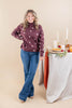 A woman stands in a studio wearing a burgundy mock-neck sweater with white polka dots and blue jeans, posed next to a styled table with candles, a pie, and autumn decorations.