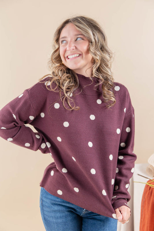 A woman wearing a maroon mock-neck long-sleeve sweater with white polka dots, paired with blue jeans, posing against a solid beige background.