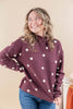 A woman wearing a maroon mock-neck long-sleeve sweater with white polka dots, paired with blue jeans, posing against a solid beige background.