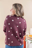 A woman shown from the back wearing a burgundy knit sweater with white polka dots, paired with blue jeans, in an indoor setting with a beige wall background.