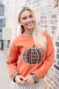 A woman wearing an orange crewneck sweatshirt with a plaid pumpkin graphic on the front, paired with cream and black pinstriped pants, standing against a brick wall.