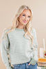 A blonde woman wearing a sage green and white patterned knit sweater with blue jeans, standing against a warm beige background.