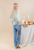 A woman with long blonde hair stands in profile wearing a sage green and white striped knit sweater and light blue jeans, standing next to a decorated table with candles, a pumpkin, and a pie.