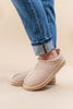 A pair of beige suede slip-on ankle boots with a braided trim around the collar, shown on a model wearing blue jeans against a solid beige background.