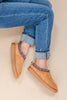 A woman wearing tan suede slip-on slippers with patterned trim, paired with cuffed blue jeans, photographed against a plain light beige background.