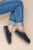 A woman wearing black suede slip-on slippers with a patterned trim and gray soles, paired with cuffed blue jeans, against a plain beige background.