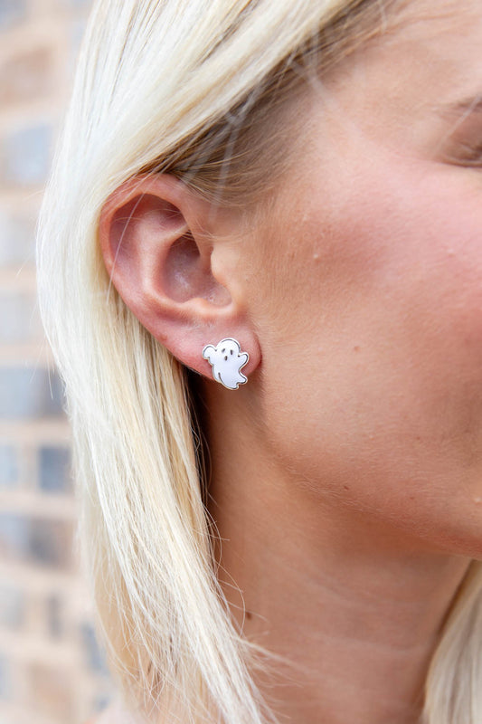 A close-up of a woman's ear wearing a small white ghost stud earring with a silver outline, set against a blurred brick wall background.