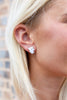 A close-up of a woman's ear wearing a small white ghost stud earring with a silver outline, set against a blurred brick wall background.