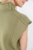 Close-up of a woman wearing a sleeveless olive green knit top with a mock neck and shoulder detailing, shown against a plain light background.