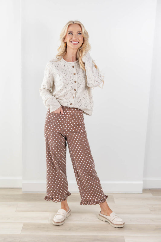 A woman modeling a cream cable-knit button-front cardigan and brown wide-leg pants with white polka dots, paired with cream loafers, standing in a bright room with a white wall and light wood floor.