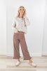 A woman modeling a cream cable-knit button-front cardigan and brown wide-leg pants with white polka dots, paired with cream loafers, standing in a bright room with a white wall and light wood floor.