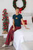 A woman sits wearing a teal blue t-shirt with a colorful Christmas wreath graphic on the chest and red plaid pajama pants.