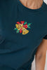 A close-up of a woman wearing a dark teal t-shirt with a colorful needlepoint holiday design on the chest featuring a red bow, green leaves, and gold bells.