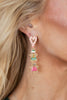 A close-up of a woman's ear wearing a gold-plated dangle earring with a pink and white striped heart-shaped top, a green and white gingerbread man charm, and a pink teddy bear charm.