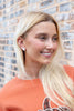 A smiling blonde woman wearing a small silver and white ghost-shaped stud earring, posing in front of a blurred brick wall background.