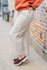 A person wearing cream wide-leg pinstriped pants with thin dark vertical stripes, paired with a rust-orange sweatshirt and brown slip-on shoes, standing outdoors next to a brick wall.