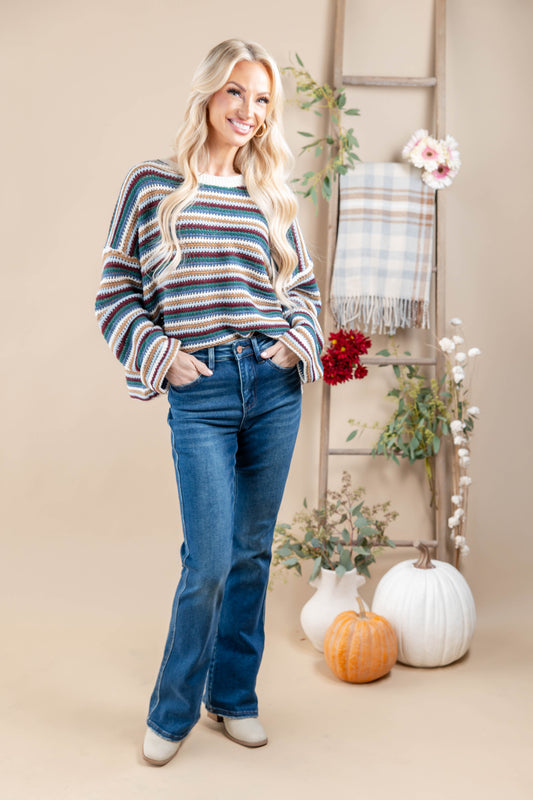 A woman stands wearing a long-sleeve knit sweater with horizontal stripes in blue, maroon, tan green, and white, paired with blue jeans and white boots. The background features a beige backdrop, a wooden ladder, a plaid throw blanket, and decorative pumpkins and flowers.