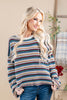 A woman with long blonde wavy hair models a cozy, long-sleeve knit sweater with horizontal stripes in teal, rust red, beige, and white. She is wearing blue jeans and standing in a warm, neutral-toned indoor setting.